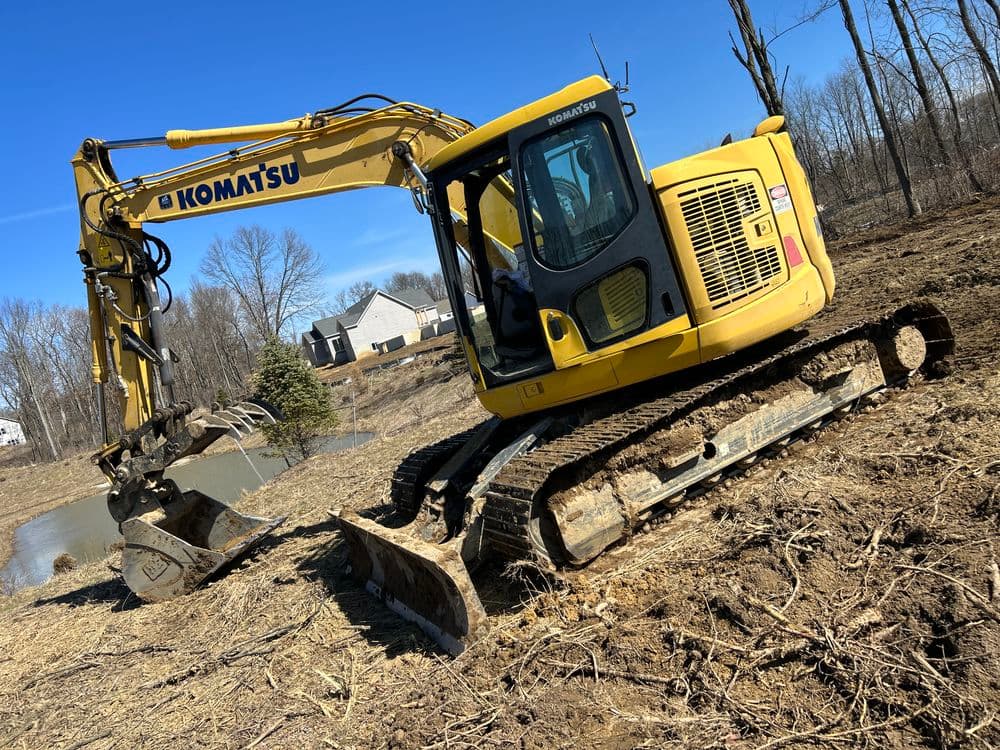 Komatsu excavator working on a construction site near a pond on a sunny day.
