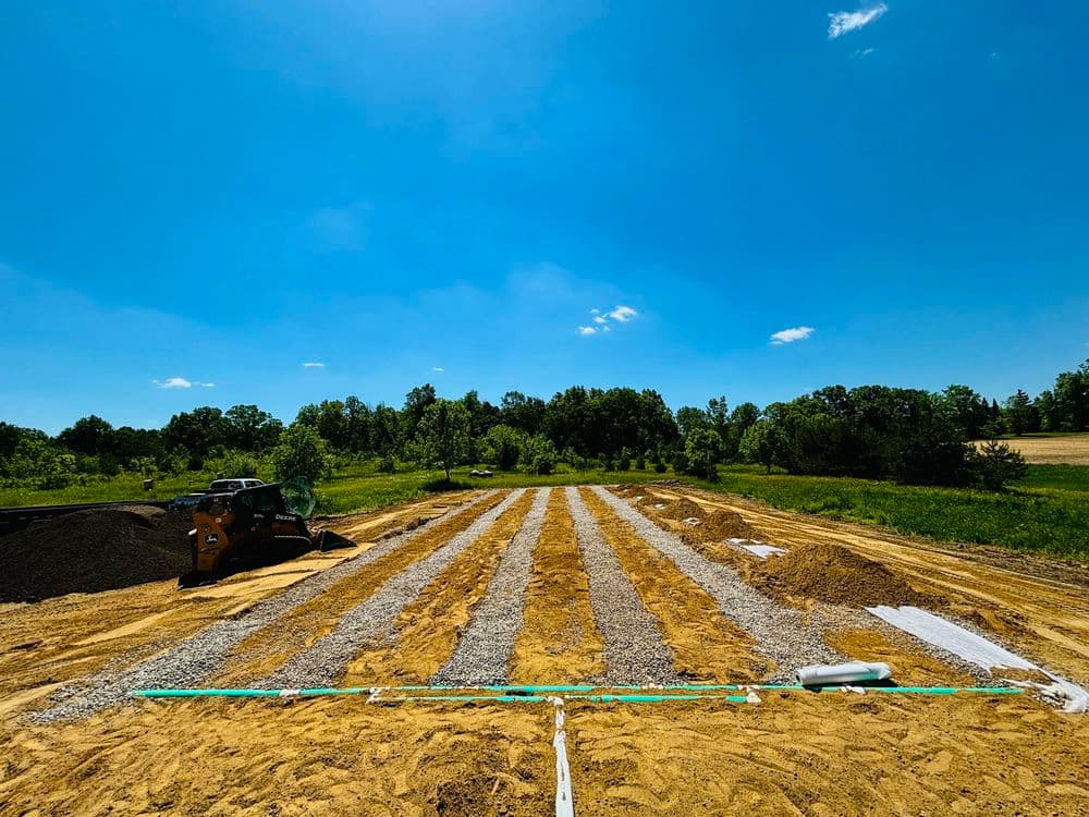 Construction site with gravel and sand layers under a clear blue sky and green trees.