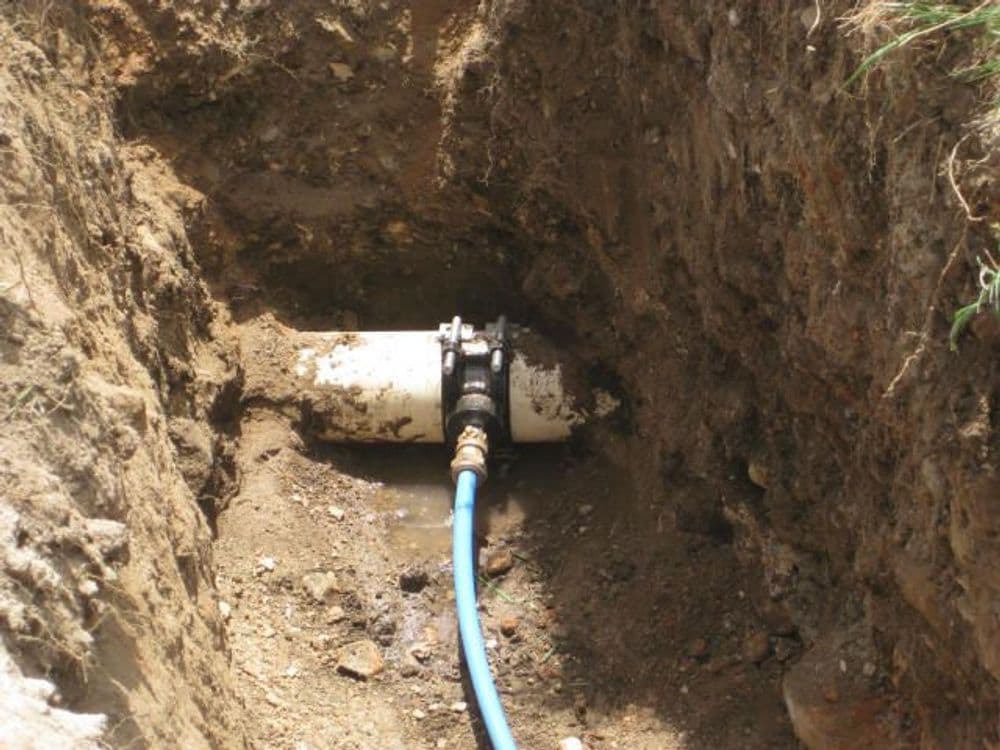 Exposed water pipe in a trench with a blue hose attached.