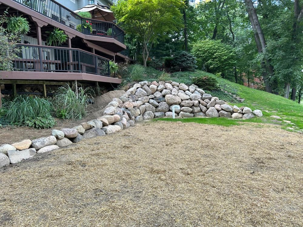 Backyard landscape featuring a stone retaining wall, grassy area, and deck in a wooded setting.